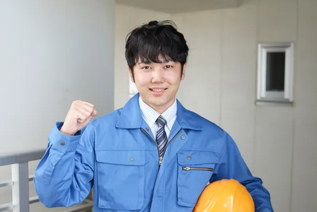 右手でガッツポーズをして左手でヘルメットを持つ青い作業着を着た男性