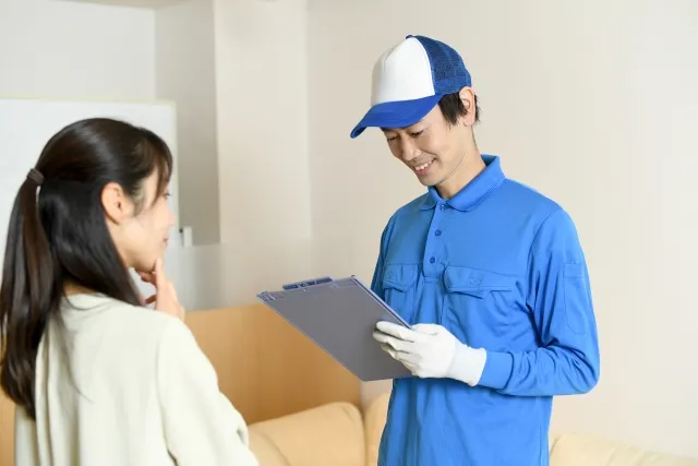 青い帽子を被って青い作業着を着た男性が女性と話しながら持っているバインダーに何かを書き込んでいる