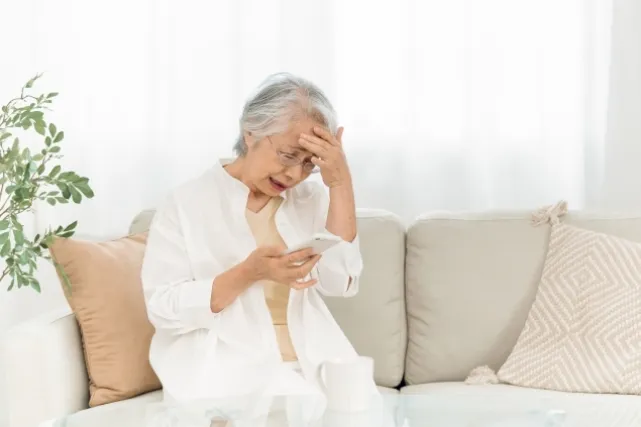 スマホを片手に困っているような表情をしている白いシャツを着た年配の女性