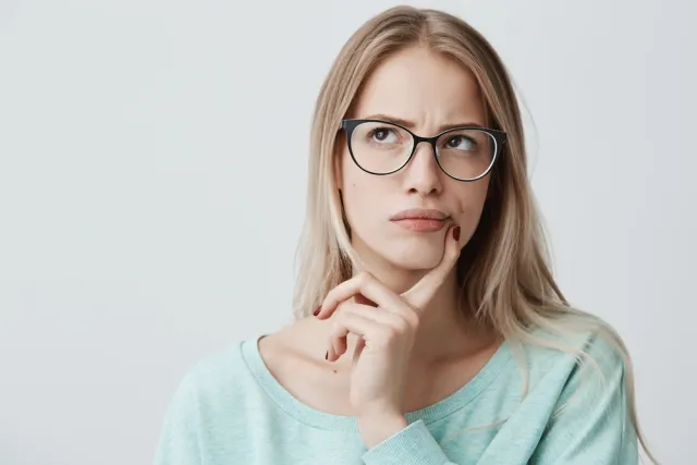唇の近くを指で押さえて何か考えている様子の薄い緑の服をきた眼鏡の外国人女性