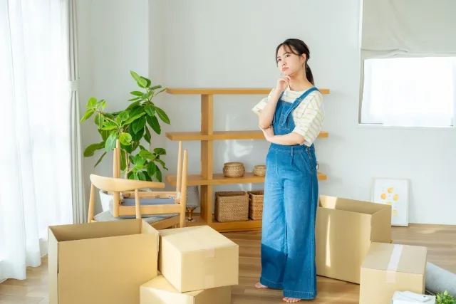 部屋でダンボールに物を入れて引っ越しの準備をしている女性が立ちながら右手を顎に当てて考えごとをしている