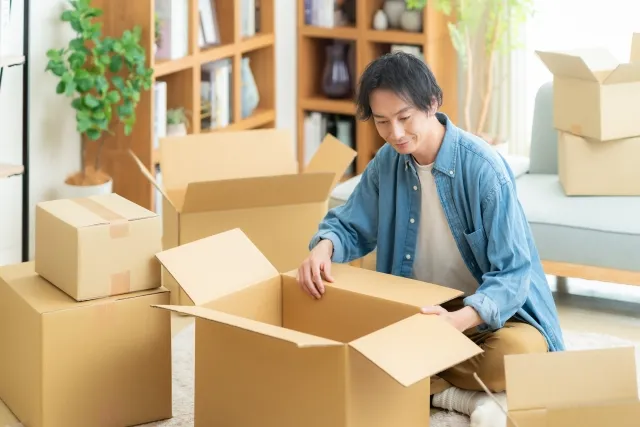 部屋で青いシャツを着た男性がダンボールに物を入れて引っ越しの準備をしている様子