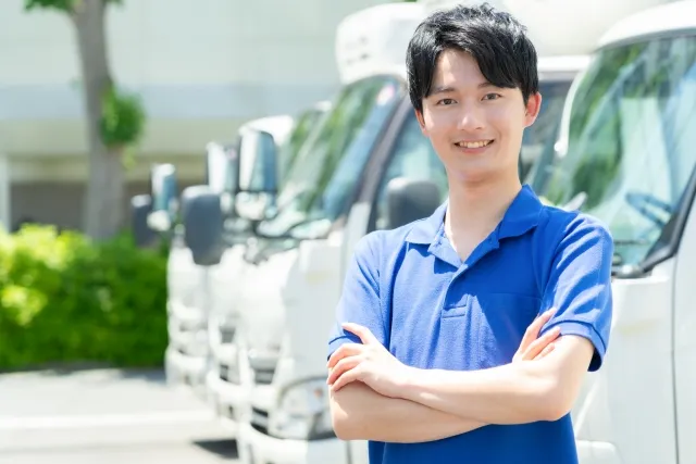 数台のトラックの前で腕組みして笑顔の引っ越し業者の青色の半そで作業着を着た男性