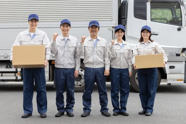 トラックの前で笑顔でガッツポーズをする白っぽい作業着を着た引っ越し業者の男性3人と女性2人