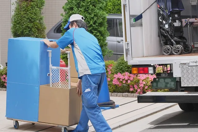トラックからダンボールに入った荷物を台車で運んでいる青い作業着を着た引っ越し業者の男性