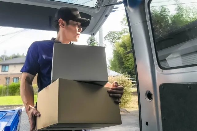 車に手に持った2つのダンボールを車の荷台に入れようとしている紺色の作業着を着た男性