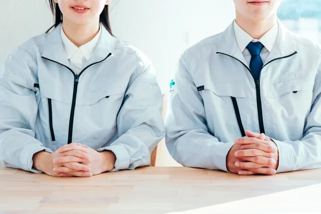 作業着を着た男女が座りながら両手を組んで机の上に置いている