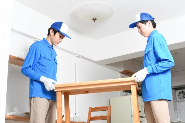 青い作業着と帽子を被った男性2人が木製のテーブルの端と端を持って運んでいる様子