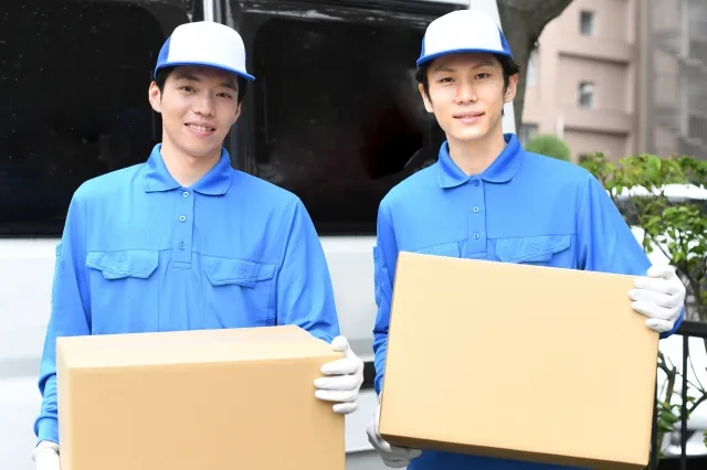 白い車の前で青い作業着と青い帽子を被ってダンボールの荷物を持つ若い男性作業員2人