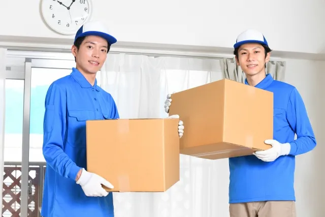 家の部屋の中で青い作業着と青い帽子を被ってダンボールの荷物を持つ若い男性作業員2人