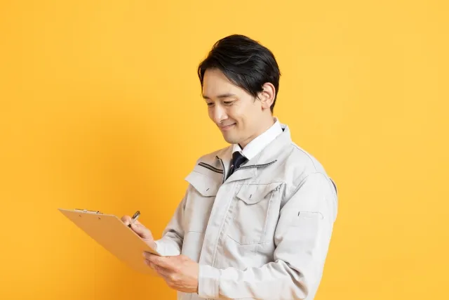 持っているバインダーに何かを書き込みながら笑顔の白っぽい作業着を着た男性作業員