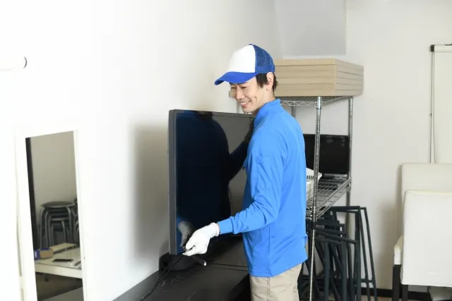 青い作業着と青い帽子を被った作業員の男性が、テレビを持ち上げて運んでいる様子