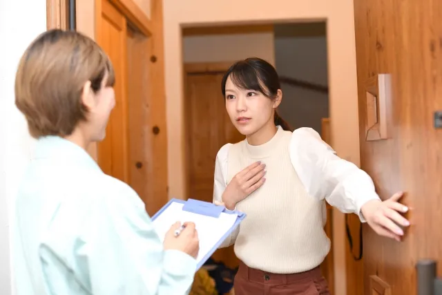 バインダーに何かを書き込もうとしている作業着の女性が訪問してきて、不安な顔をしている応対している女性