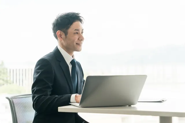 スーツを着た男性が椅子に座りテーブルに置いたノートパソコンを操作している