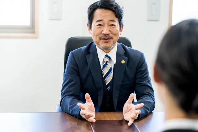 スーツを着た中年の男性が椅子に座りテーブル越しの女性に話しかけている