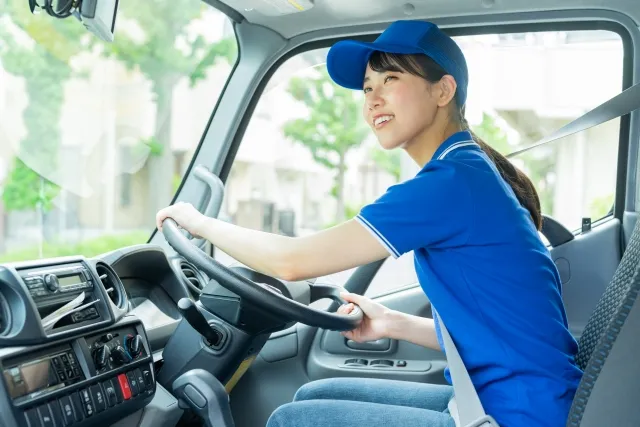 青い半そでのポロシャツと青い帽子を被った笑顔の女性がトラックを運転している