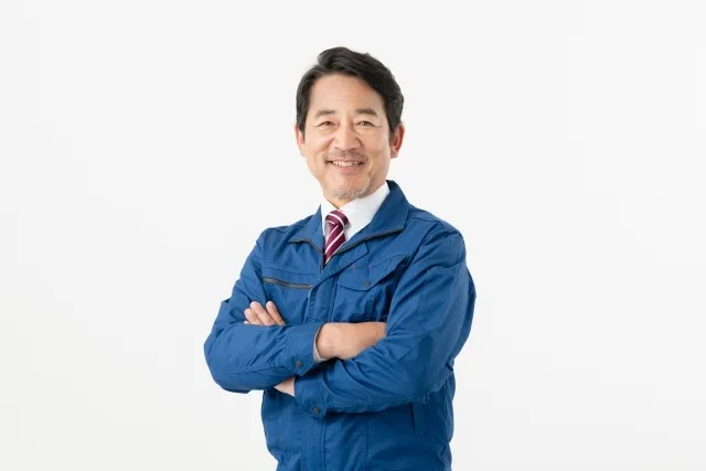 青い作業着を着た年配の男性が腕を組みながら微笑んでいる
