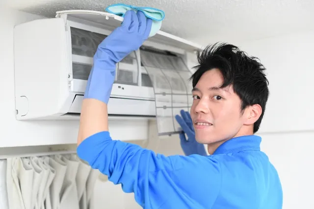 エアコンのカバーを開けてフィルターを外そうとしている青い作業着の男性