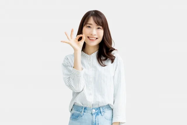 右手でOKサインをしながら笑顔を見せるストライプのシャツとジーンズ姿の女性